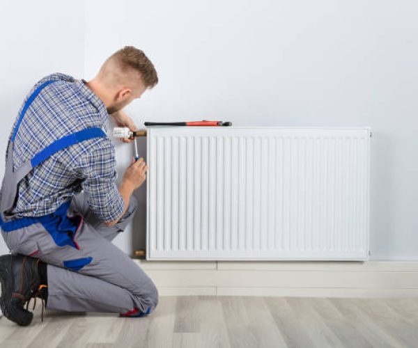 Rear View Of Male Plumber Fixing Thermostat With Screwdriver And Wrench On Radiator